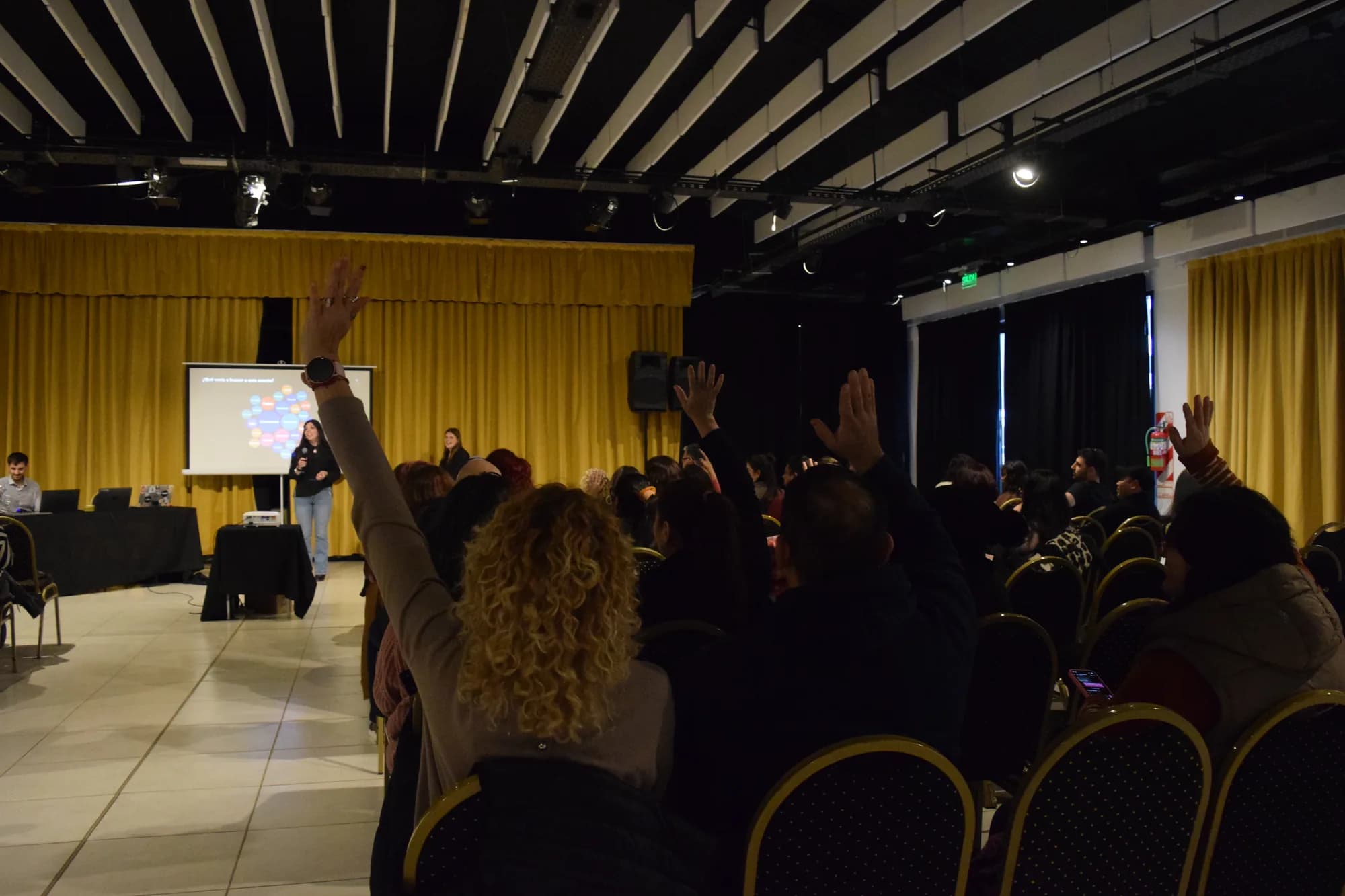 Desde el fondo de la sala, el público levanta la mano frente al escenario