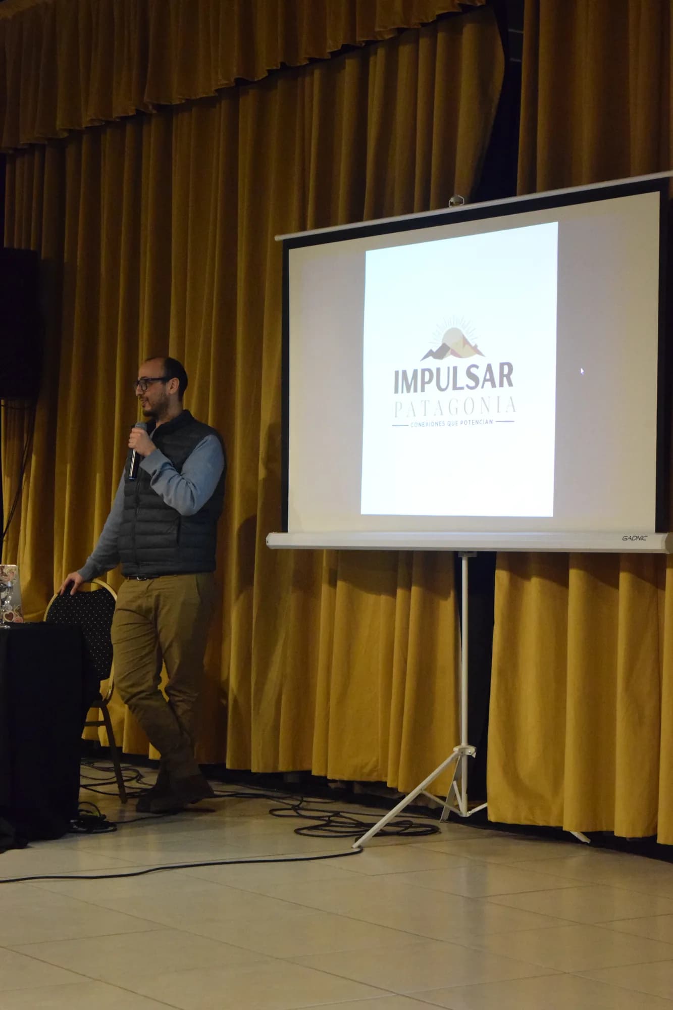 Orador con micrófono junto a la pantalla con el logo de Impulsar